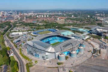 Aerial view of Etihad Stadium in Manchester, surrounded by parking lots, training grounds, and city buildings in the distance.
