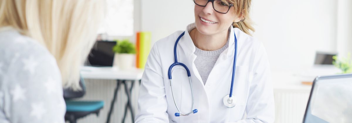 Smiling doctor talking with a patient in an office with a laptop and stethoscope on the desk.